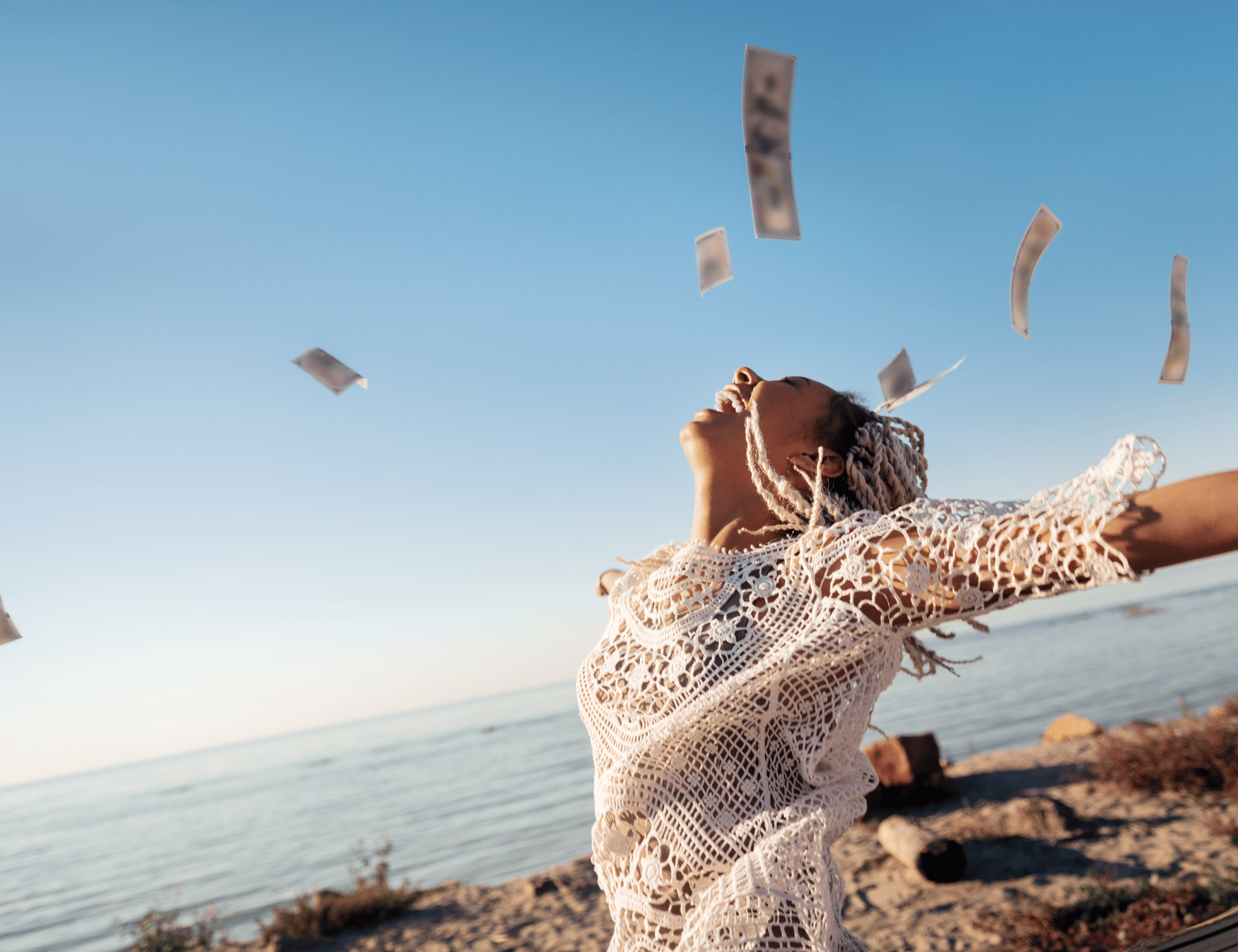 Jeune femme souriante en robe blanche en dentelle, les bras ouverts sur une plage, avec des billets de banque flottant dans les airs sous un ciel bleu