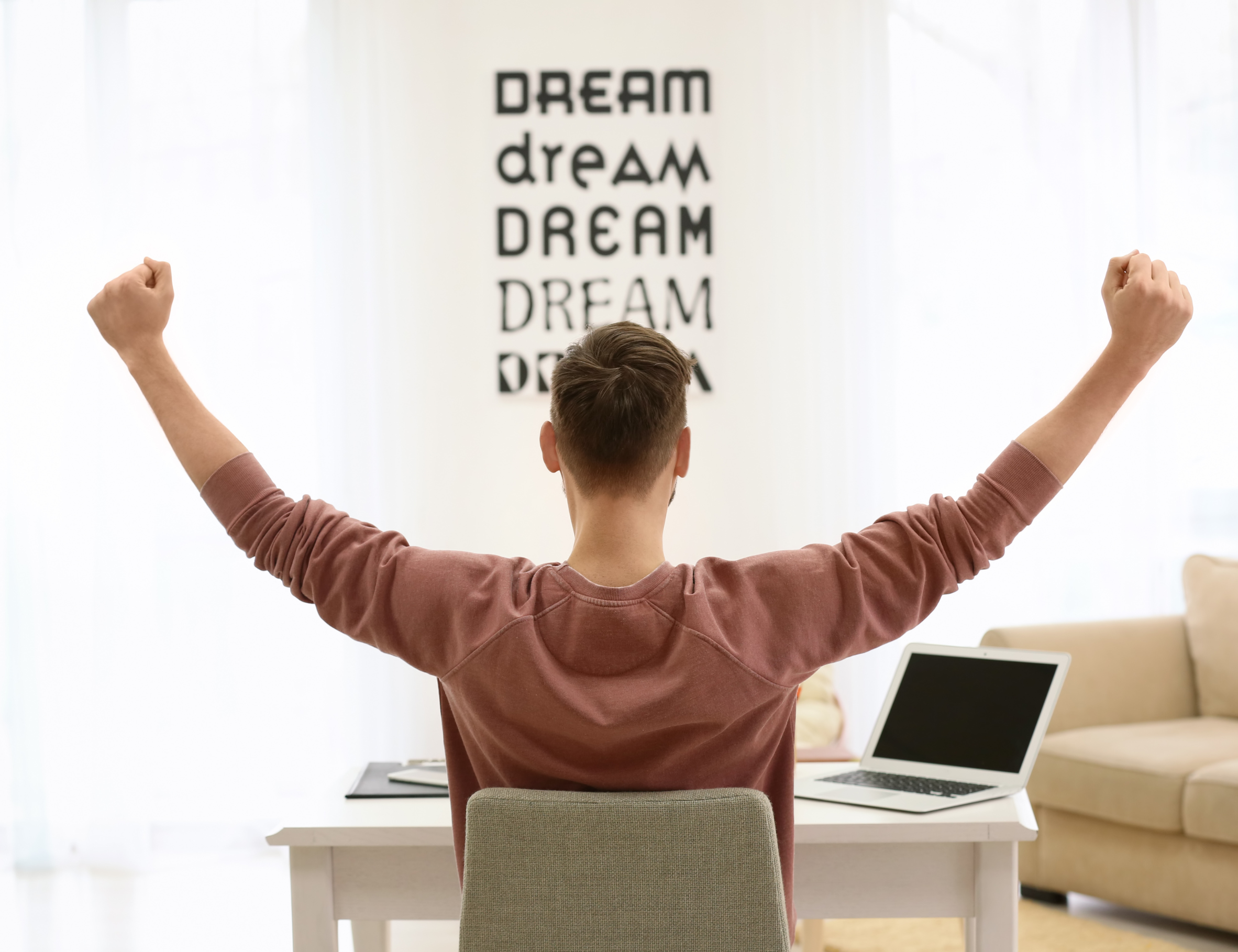 Jeune entrepreneur levant les bras en signe de victoire devant son ordinateur, dans un espace de travail inspirant avec le mot DREAM affiché sur le mur